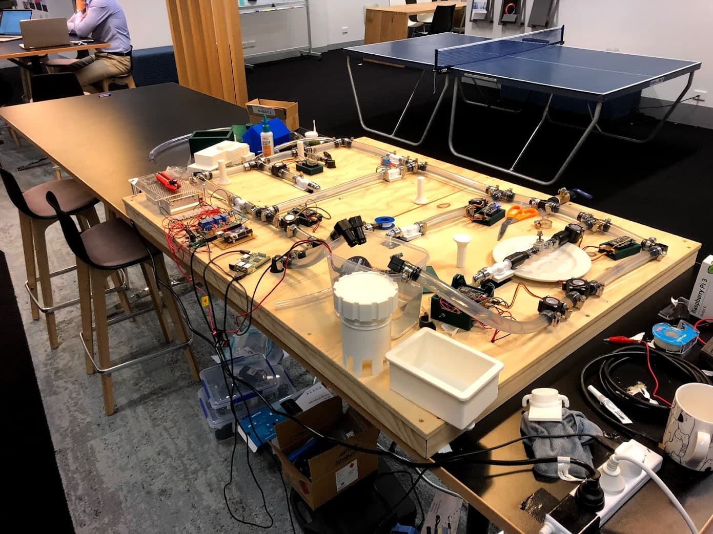 The exposed H2.0 water loop during team integration, with pipes, valves, connected components, tools and cables across the timber base.